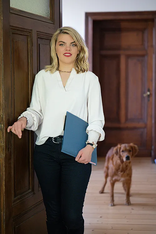 Laura Rostohar, Mitarbeiterin am Empfang, steht an einer geöffneten Tür und lächelt in die Kamera. Im Hintergrund ein freundlich blickender Hund.