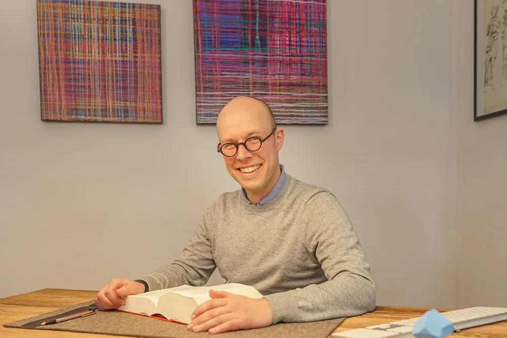 Rechtsanwalt und Notar Dr.jur. Tim Illner sitzt hinter einem Schreibtisch, mit einem aufgeschlagenen Buch vor sich, und schaut lächelnd in die Kamera.